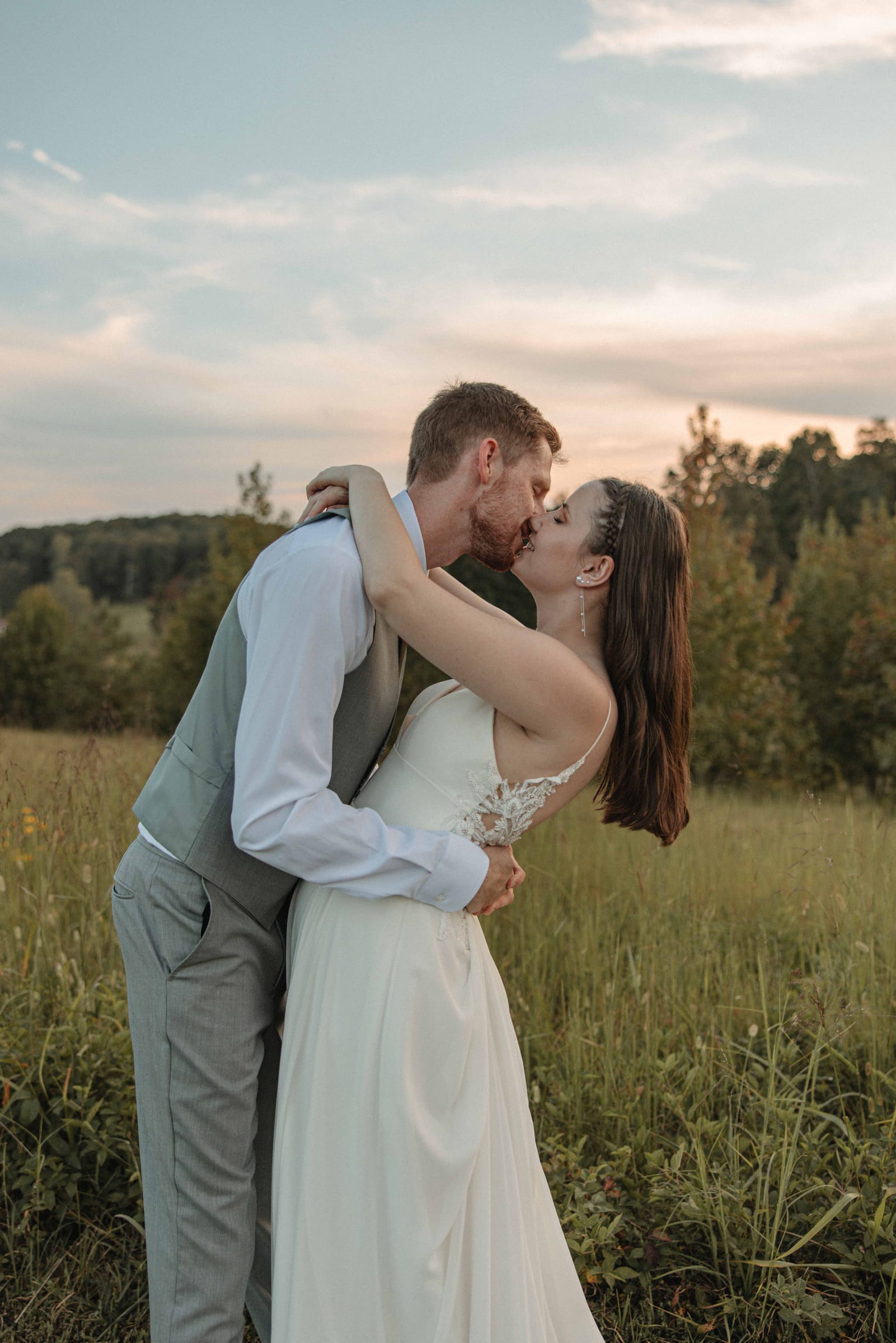 Bride and groom sharing a romantic kiss in the tall grass meadow at Cedar Hills Farm at dusk with autumn trees in the background