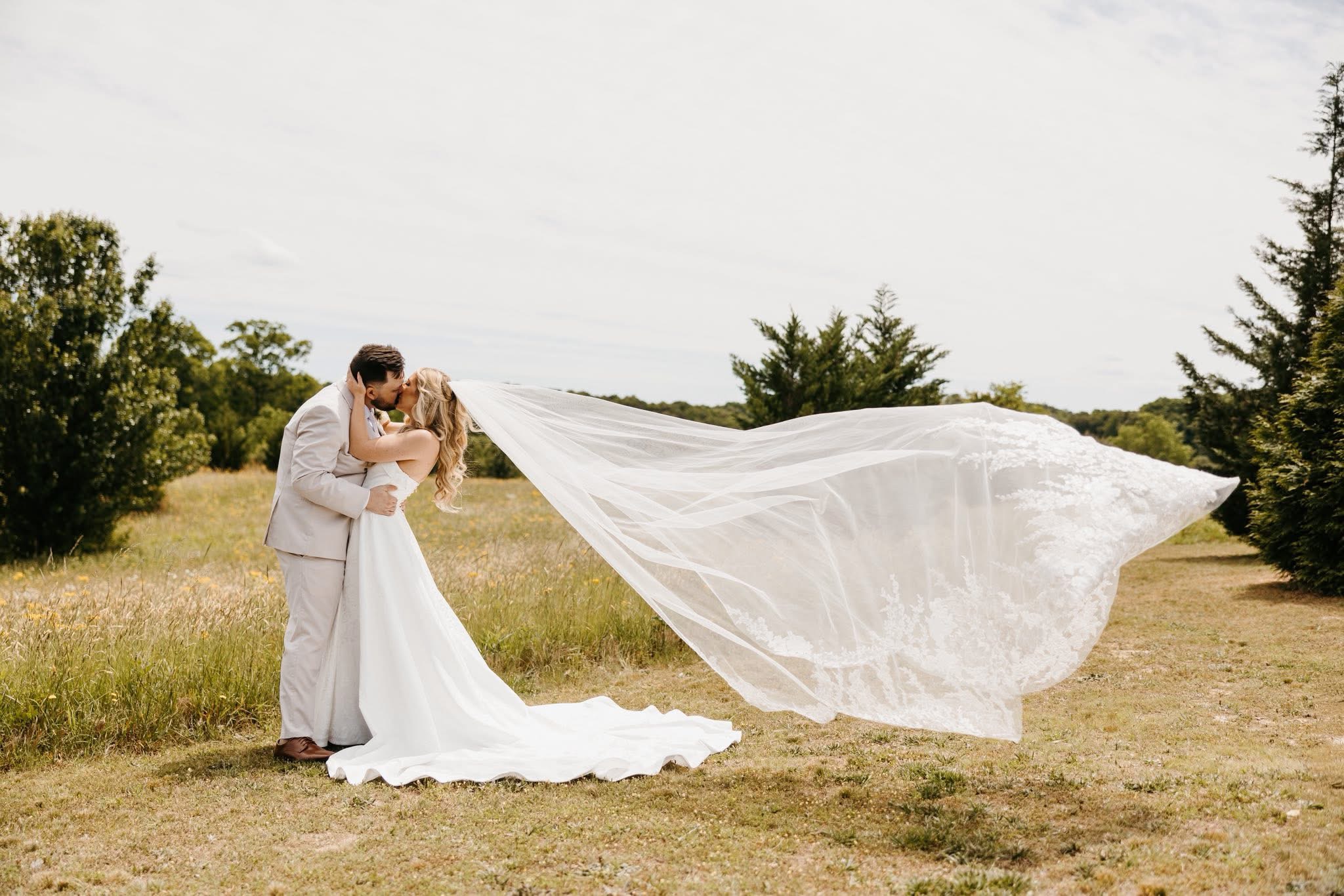 Bride and groom kissing in the open meadow at Cedar Hills Farm with the cathedral veil billowing dramatically in the wind