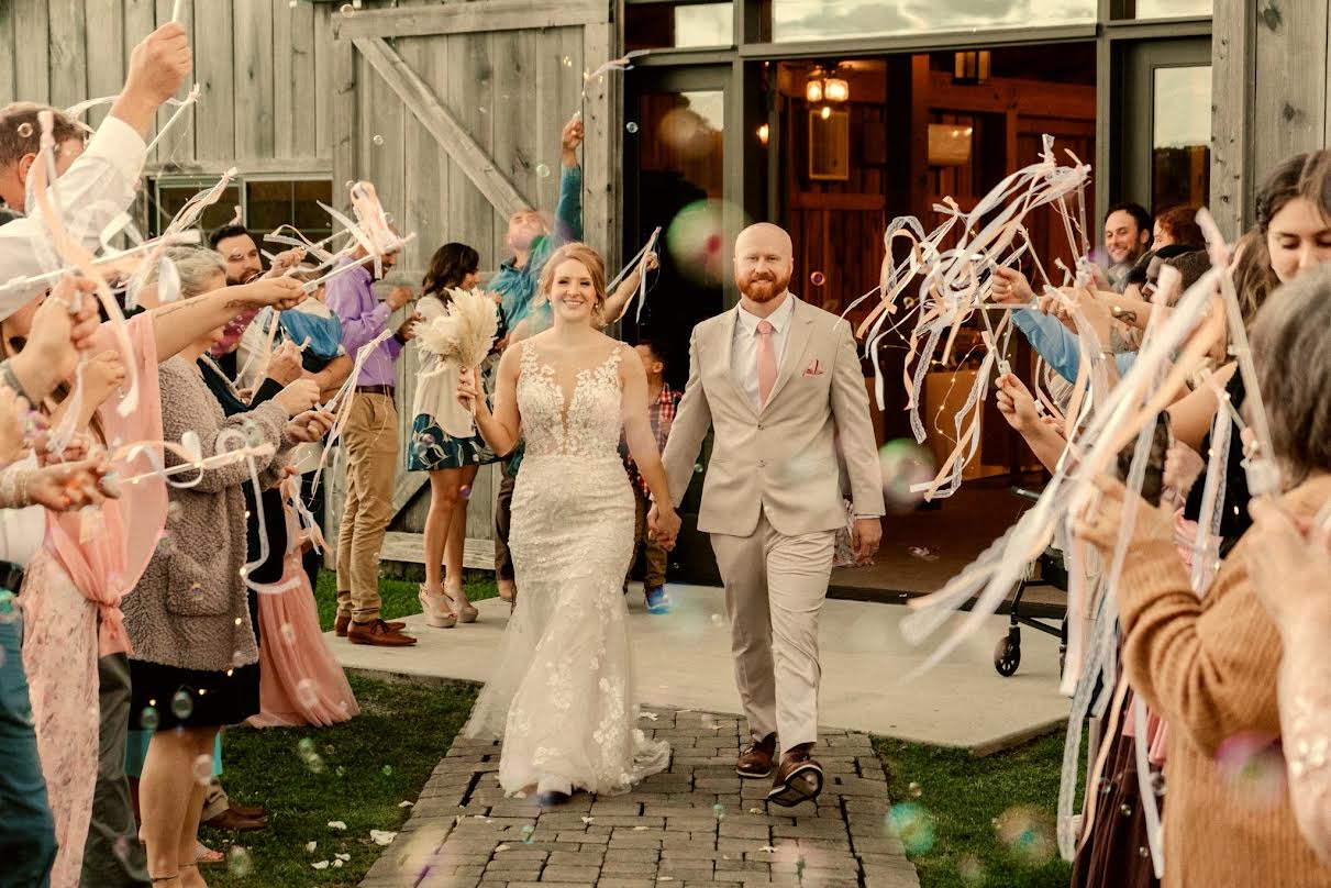Bride and groom walking through a ribbon-wand send-off with guests cheering outside the Cedar Hills Farm reception barn