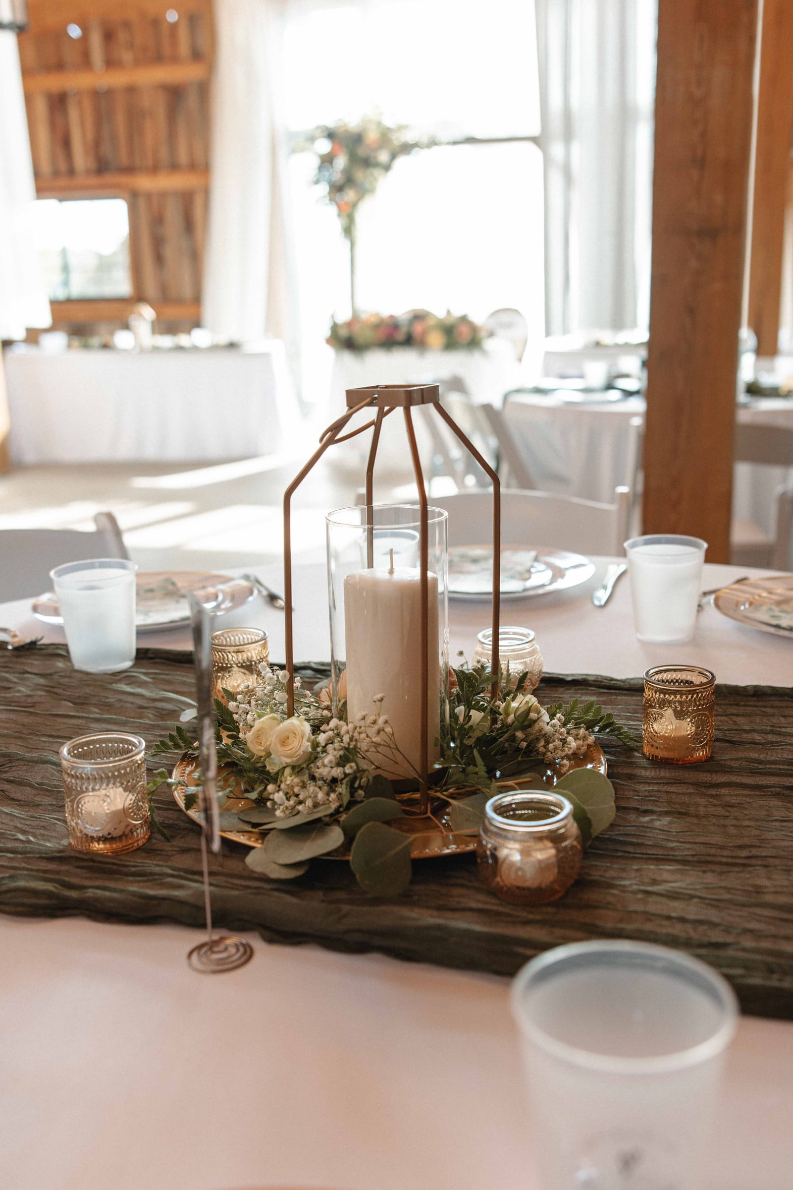 Rustic wedding table centerpiece at Cedar Hills Farm with a pillar candle in a geometric metal lantern surrounded by baby's breath, eucalyptus, and gold mercury glass votives on a wood slab