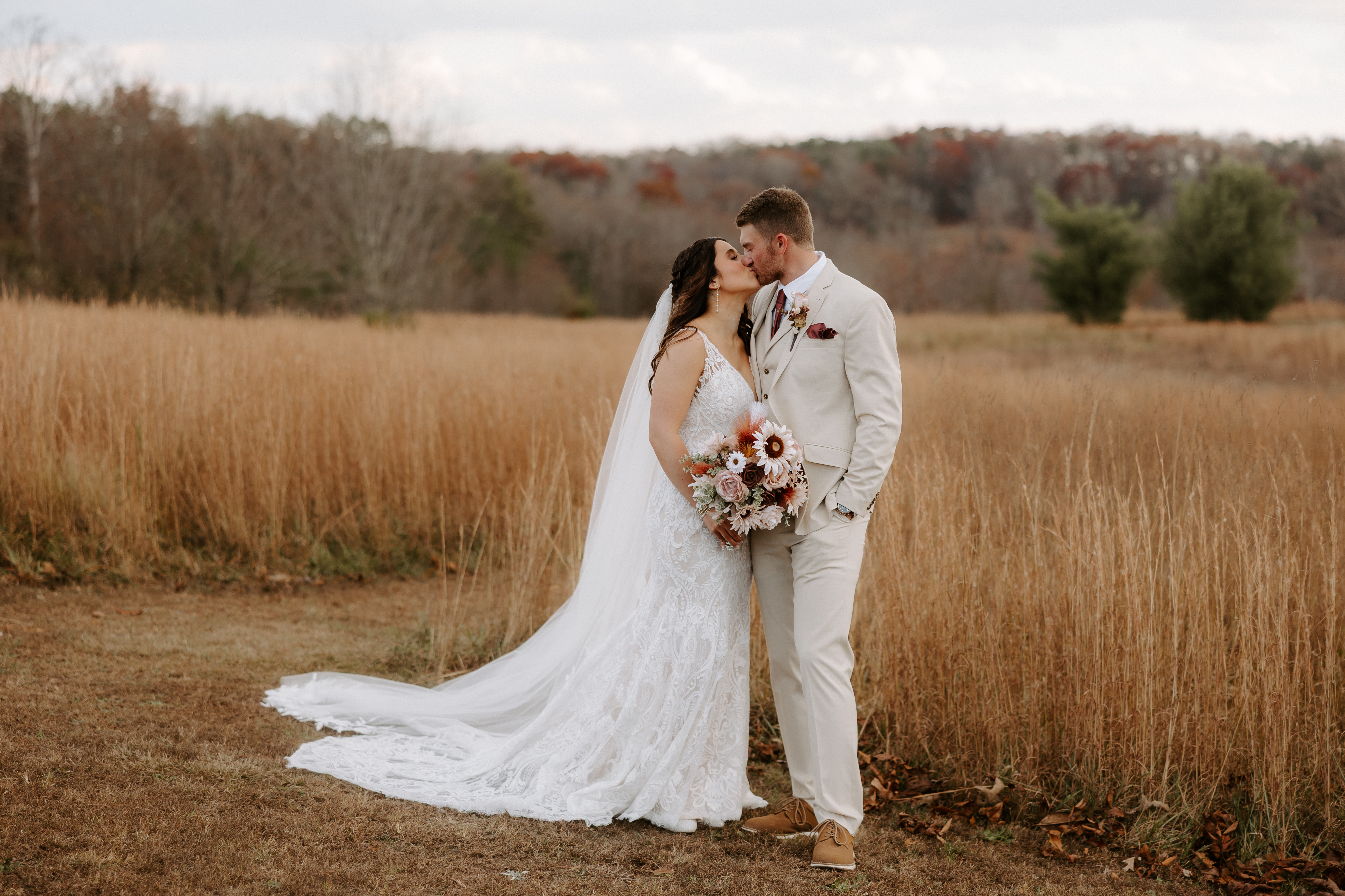 Bride and groom kissing in the tall golden grass meadow at Cedar Hills Farm in late autumn with fall foliage in the background, bride in a lace gown holding a sunflower and burgundy bouquet