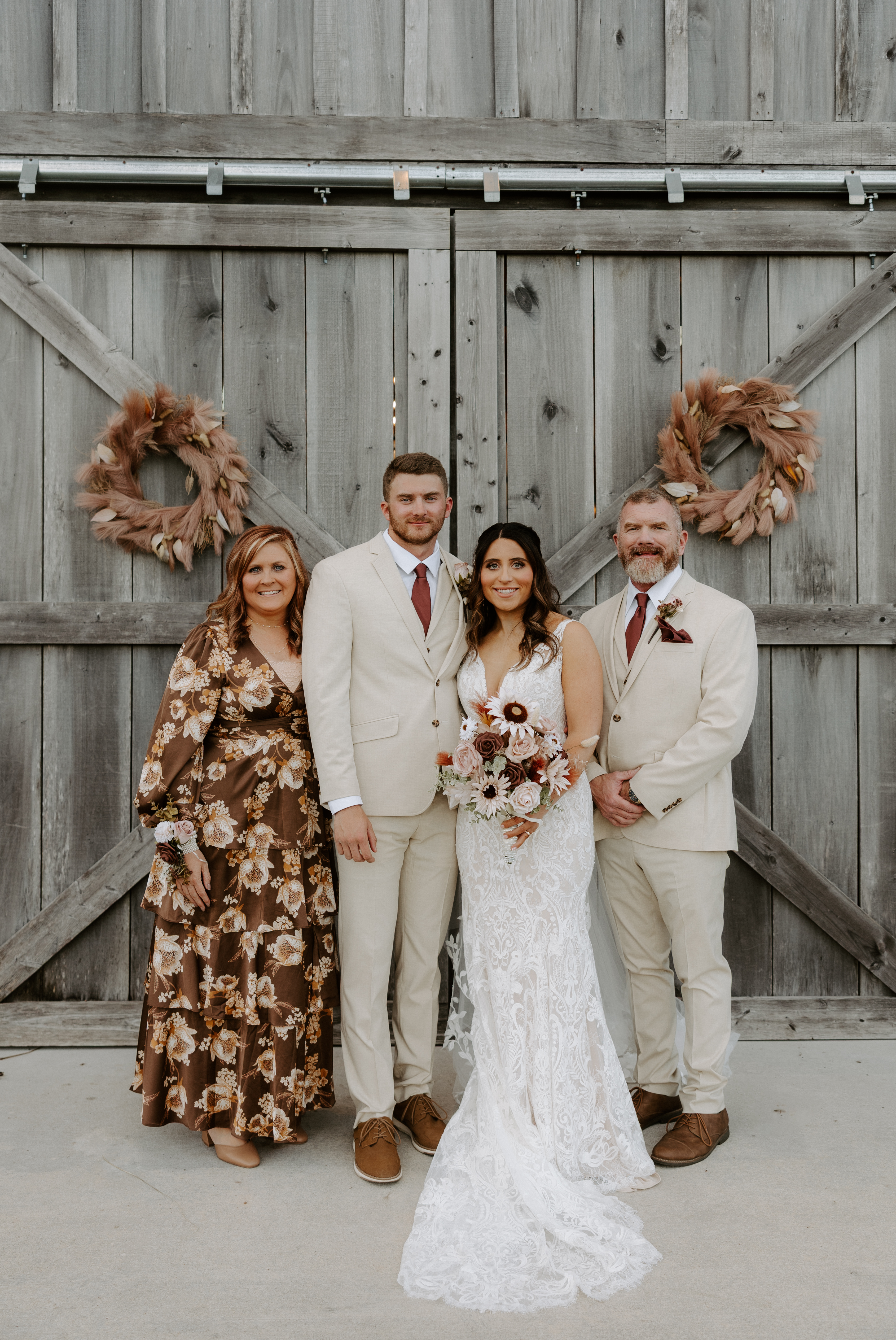 Bride and groom posing with his parents in front of the Cedar Hills Farm barn doors decorated with dried floral wreaths, groom in a khaki suit and bride holding a rust and white bouquet