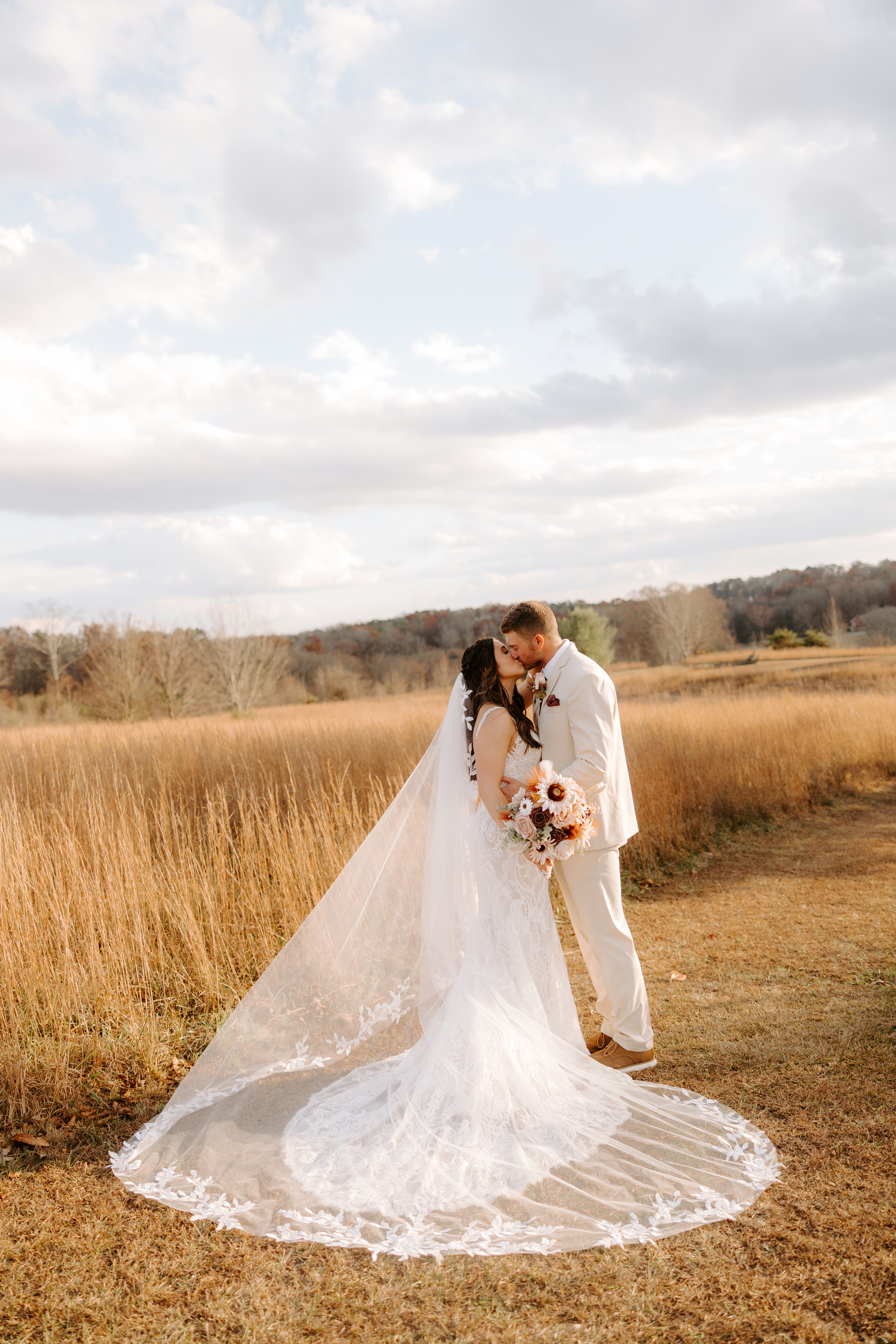 Bride and groom kissing in the open golden meadow at Cedar Hills Farm with the bride's cathedral veil spread dramatically across the grass under a dramatic cloudy sky
