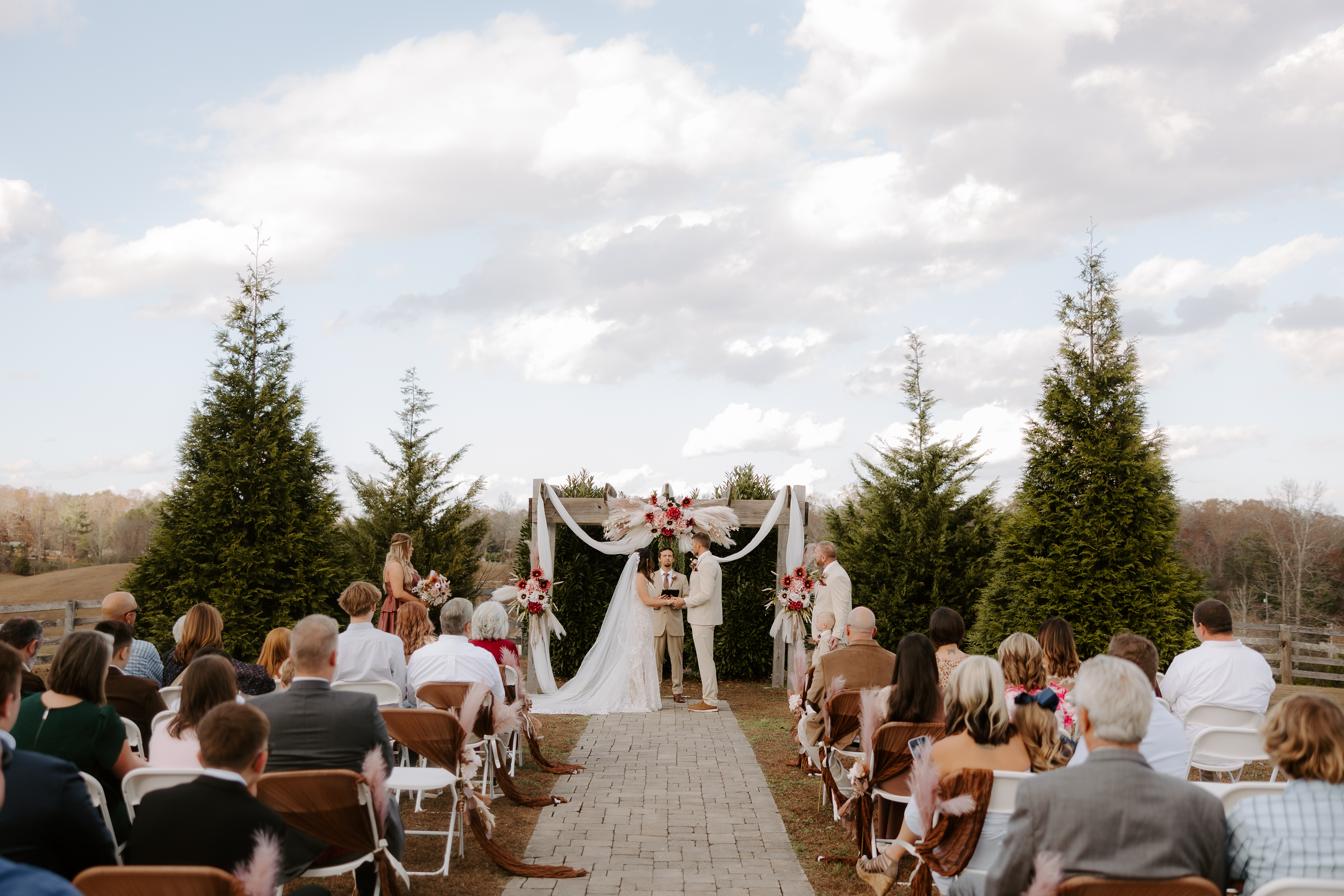 Outdoor wedding ceremony in progress at Cedar Hills Farm with guests seated, bride and groom at the altar under a white-draped arch with red floral accents, flanked by evergreen trees