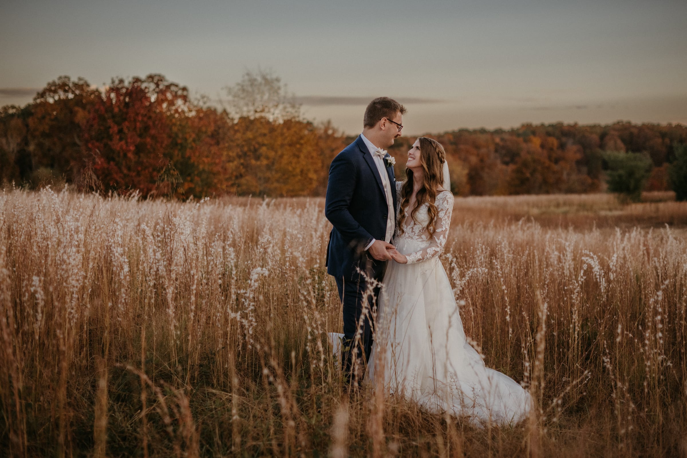 Bride and groom standing together in the tall grass meadow at Cedar Hills Farm surrounded by fall foliage at dusk
