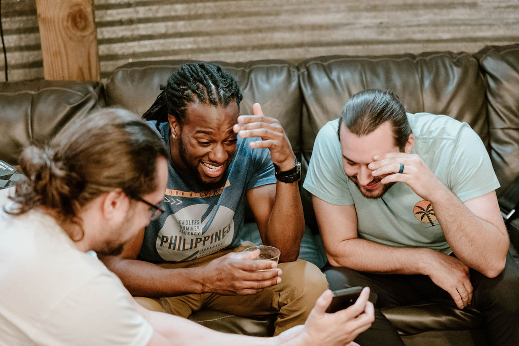 Groomsmen laughing together in casual clothes on the leather couch in the groom's suite at Cedar Hills Farm before getting dressed