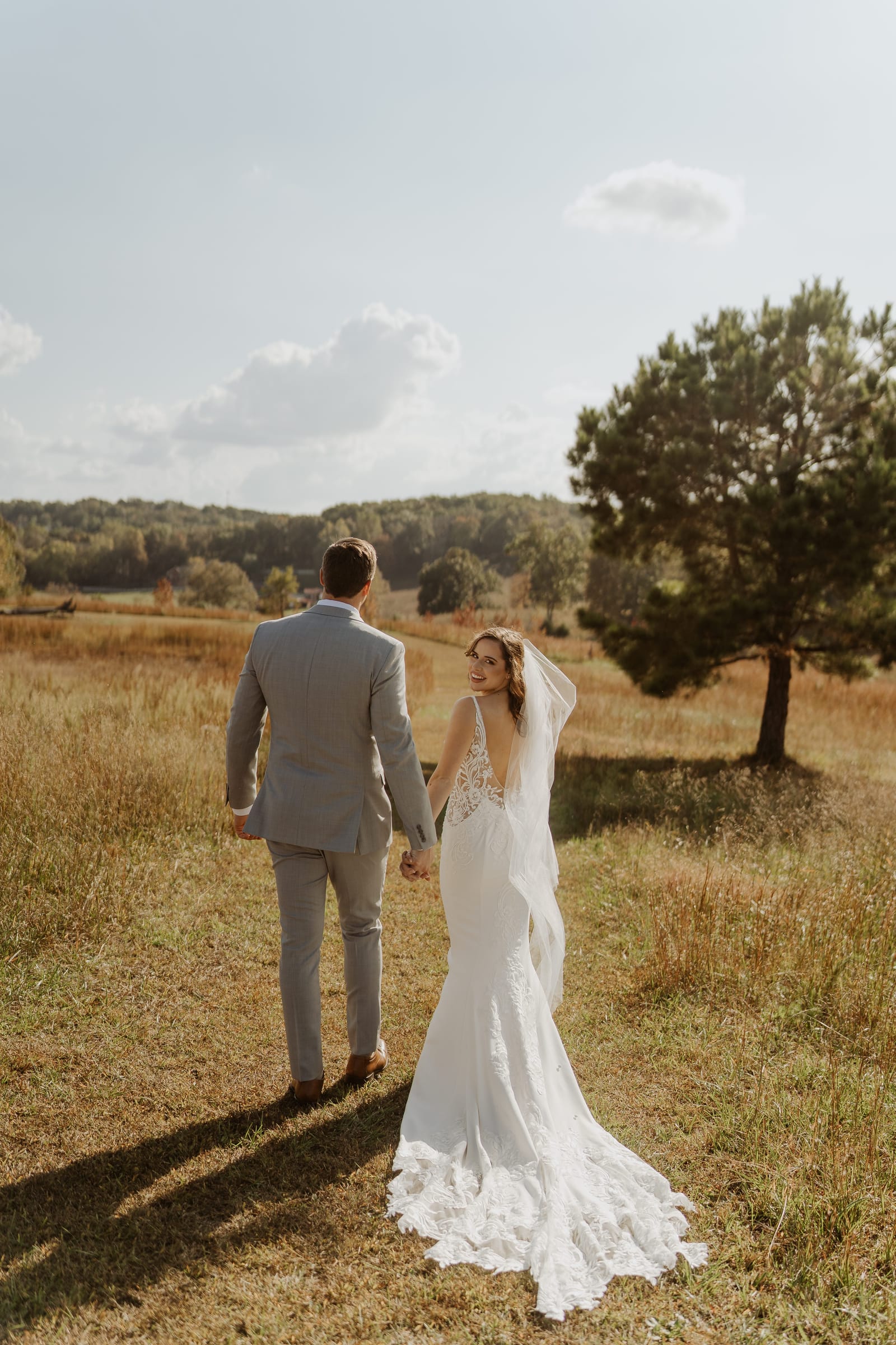 Bride in a backless lace gown glancing back over her shoulder as the couple walks hand-in-hand through the golden autumn meadow at Cedar Hills Farm