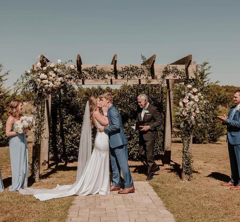 Bride and groom sharing their first kiss under the cedar ceremony arch at Cedar Hills Farm with the maid of honor and officiant visible