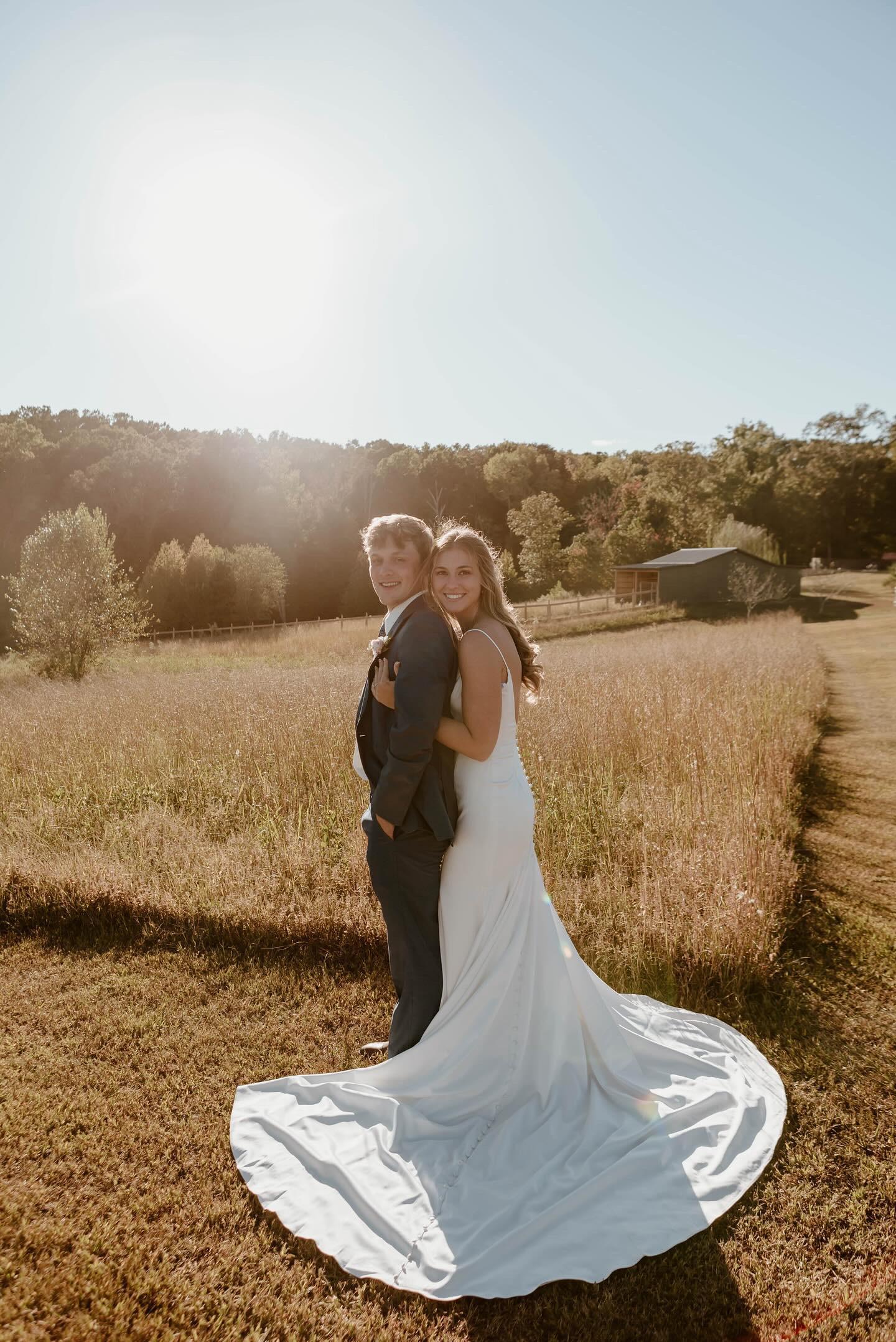 Bride and groom standing back to back in the golden sunlit meadow at Cedar Hills Farm smiling over their shoulders with the barn visible in the background