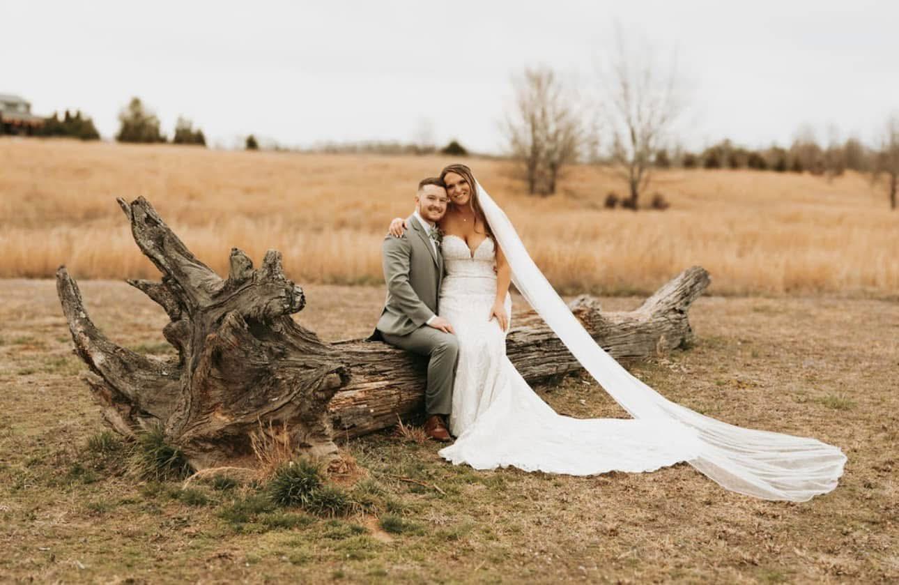 Bride and groom sitting together on the driftwood log in the open meadow at Cedar Hills Farm with the veil spread across the grass