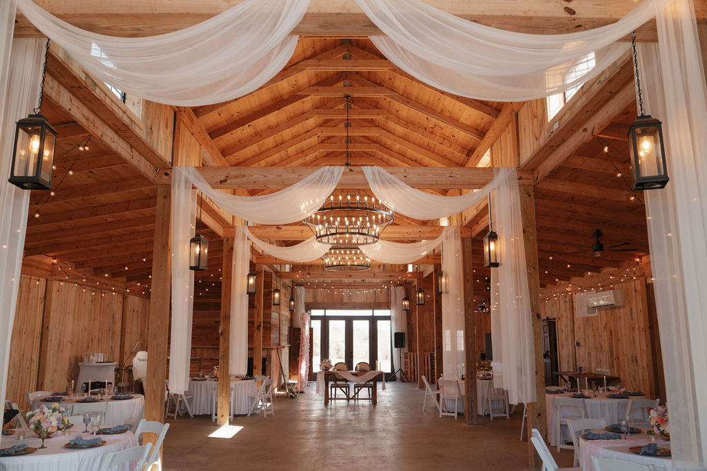 Cedar Hills Farm reception barn interior looking toward the entrance doors with white fabric draping, ring chandeliers, string lights, and round tables set for a spring wedding