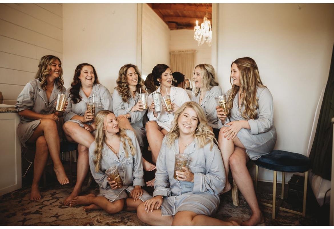 Bride and bridesmaids in matching light grey pajama sets holding iced drinks and laughing together in the bridal suite at Cedar Hills Farm