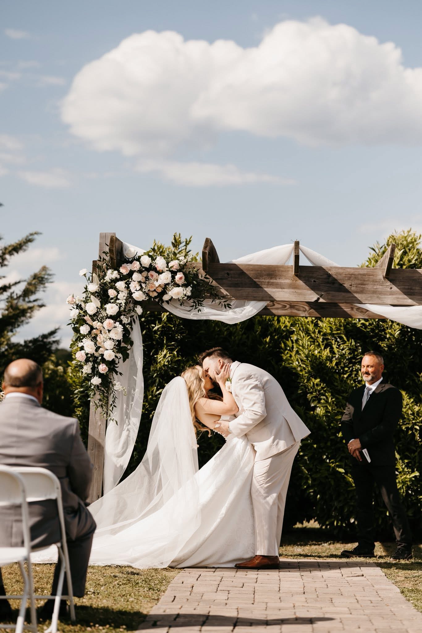 Groom in an ivory suit dipping his bride for their first kiss under the cedar ceremony arch at Cedar Hills Farm with blush roses and white draping
