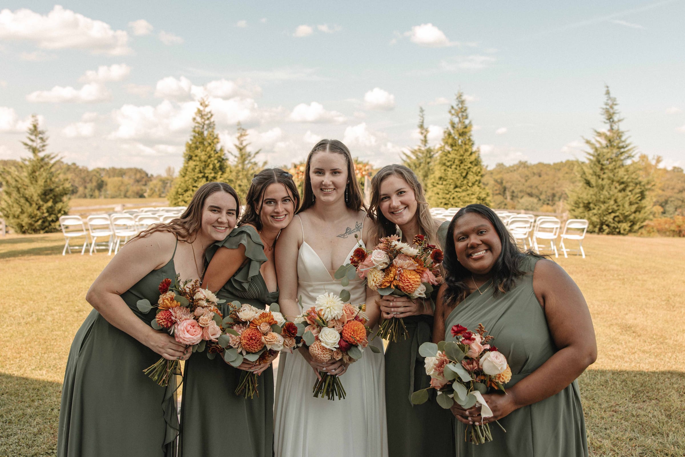 Bride with four bridesmaids in sage green dresses holding fall bouquets on the ceremony lawn at Cedar Hills Farm with chairs set in the background