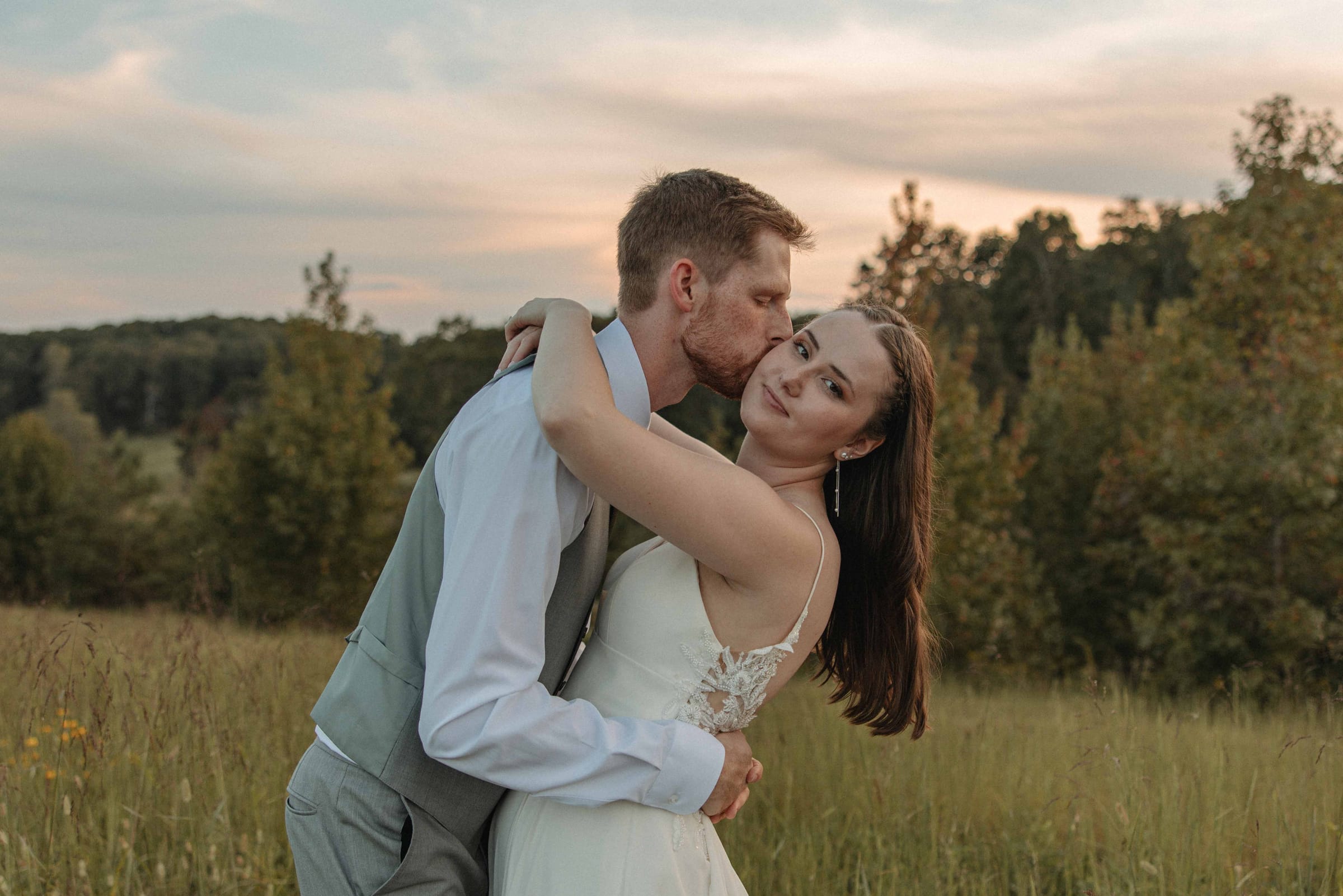 Groom kissing his bride on the cheek as she smiles at the camera during golden hour couple portraits in the tall grass meadow at Cedar Hills Farm